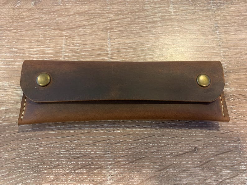 May include: A rectangular brown leather case with a fold-over flap secured by two brass-colored metal snap buttons. The leather features visible stitching along the side edges and a smooth, matte finish with natural variations in the brown tone.