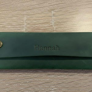May include: A rectangular dark green leather pencil case featuring a flap closure secured by two brass-toned metal snaps. The name Hannah is embossed in the center of the flap, and the edges are stitched with matching green thread.