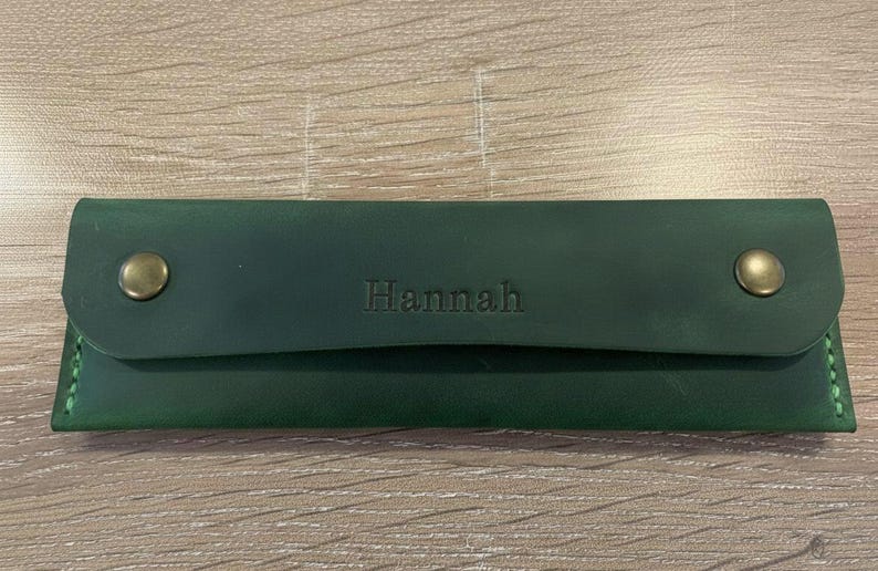 May include: A rectangular dark green leather pencil case featuring a flap closure secured by two brass-toned metal snaps. The name Hannah is embossed in the center of the flap, and the edges are stitched with matching green thread.