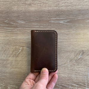 Puede incluir: Un tarjetero rectangular de cuero marr&oacute;n oscuro con esquinas redondeadas y detalles de costuras gruesas de color claro a lo largo de los bordes. El cuero tiene una textura mate y suave con grano natural visible y variaciones de tono.