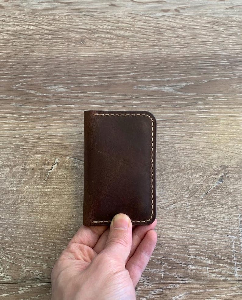 Puede incluir: Un tarjetero rectangular de cuero marr&oacute;n oscuro con esquinas redondeadas y detalles de costuras gruesas de color claro a lo largo de los bordes. El cuero tiene una textura mate y suave con grano natural visible y variaciones de tono.