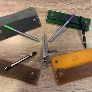 May include: Four rectangular leather pen cases in charcoal, green, dark brown, and tan, each featuring brass snap closures and white stitching. The tan case is open, revealing a LEE logo. Included are three pens, a drafting compass, and a divider.