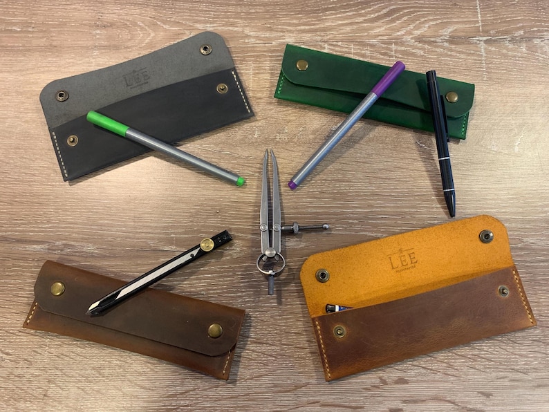 May include: Four rectangular leather pen cases in charcoal, green, dark brown, and tan, each featuring brass snap closures and white stitching. The tan case is open, revealing a LEE logo. Included are three pens, a drafting compass, and a divider.