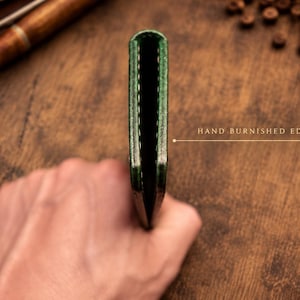 Puede incluir: Una cartera de cuero verde delgada vista de lado, que muestra una construcci&oacute;n plegada con costuras visibles a lo largo del borde. El texto "HAND BURNISHED EDGES" est&aacute; impreso junto a una l&iacute;nea que se&ntilde;ala el borde liso y acabado del cuero.