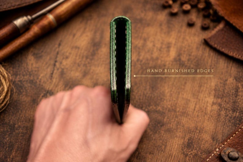 Puede incluir: Una cartera de cuero verde delgada vista de lado, que muestra una construcci&oacute;n plegada con costuras visibles a lo largo del borde. El texto "HAND BURNISHED EDGES" est&aacute; impreso junto a una l&iacute;nea que se&ntilde;ala el borde liso y acabado del cuero.