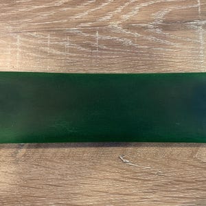 May include: A rectangular dark green leather pen case featuring a smooth finish and vibrant green stitching along the two short sides. The case has a flat, elongated shape with clean edges and a consistent, deep forest green color throughout the surface.