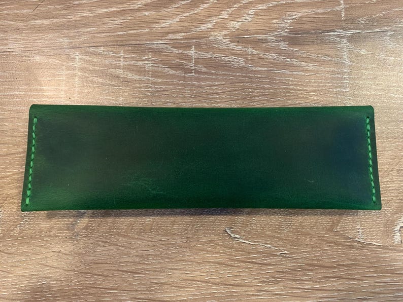 May include: A rectangular dark green leather pen case featuring a smooth finish and vibrant green stitching along the two short sides. The case has a flat, elongated shape with clean edges and a consistent, deep forest green color throughout the surface.