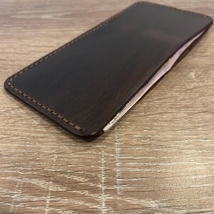 May include: A dark brown leather wallet with a rectangular shape. The edges are stitched with a lighter brown thread. The wallet appears to be empty, with a glimpse of a light-colored interior. The wallet is resting on a wooden surface.