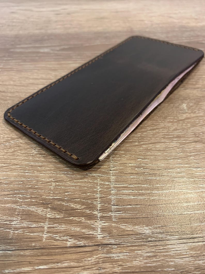 May include: A dark brown leather wallet with a rectangular shape. The edges are stitched with a lighter brown thread. The wallet appears to be empty, with a glimpse of a light-colored interior. The wallet is resting on a wooden surface.