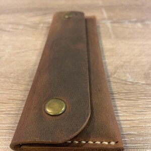May include: A rectangular brown leather case with a fold-over flap secured by a single brass-colored metal snap button. The edges of the case feature visible light-colored stitching along the bottom seam, showcasing a smooth, matte texture.
