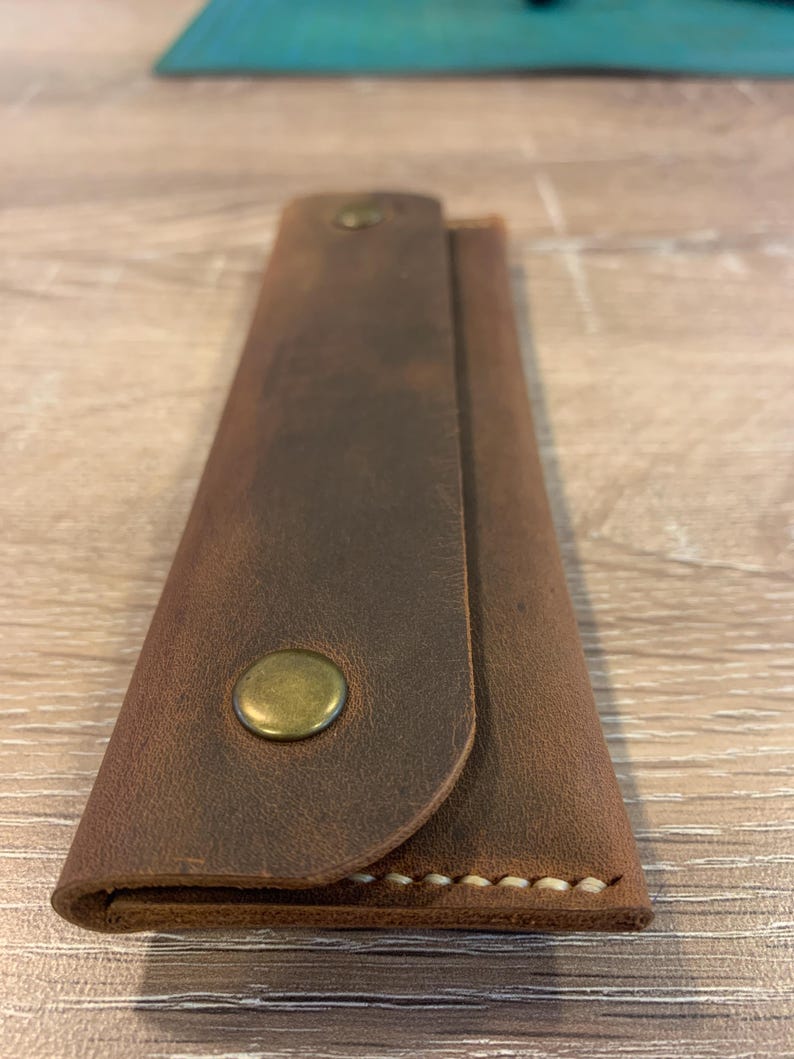 May include: A rectangular brown leather case with a fold-over flap secured by a single brass-colored metal snap button. The edges of the case feature visible light-colored stitching along the bottom seam, showcasing a smooth, matte texture.