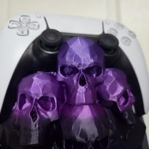 May include: A white and black video game controller rests on a purple skull-themed stand. The stand features three skulls with a faceted design, creating a gothic aesthetic. The controller's buttons and joysticks are visible.
