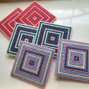 Puede incluir: Un conjunto de posavasos cuadrados hechos a mano en varios colores. Los posavasos presentan un patrón geométrico de diamantes concéntricos en tonos de rojo, azul, rosa y amarillo. Cada posavasos mide aproximadamente 10 cm cuadrados.