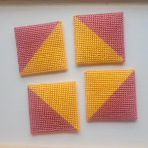May include: Four square coasters with a diagonal color block design. Each coaster is divided into two triangles, one in a dusty rose color and the other in a golden yellow. The coasters are made of a textured, woven material.