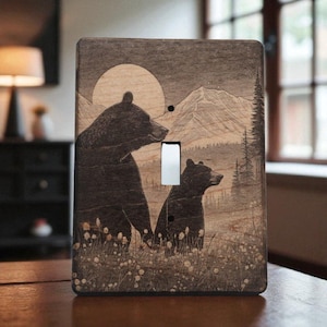 May include: Wooden light switch cover featuring a detailed illustration of two bears in a mountain landscape. The design includes a large sun, mountains, and a forest, with a light switch opening in the center.