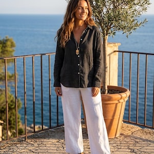 Puede incluir: Una camisa de manga larga gris carbón con botones, cuello y botones blancos, combinada con pantalones blancos holgados. Un collar con colgante dorado cuelga de una cadena larga y se llevan sandalias de tiras de color marrón.