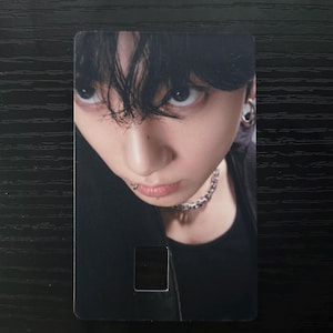 May include: A rectangular card featuring a close-up portrait of a person with dark hair and a piercing. The person is wearing a black top and a silver chain necklace. The card has a small rectangular cutout near the bottom.