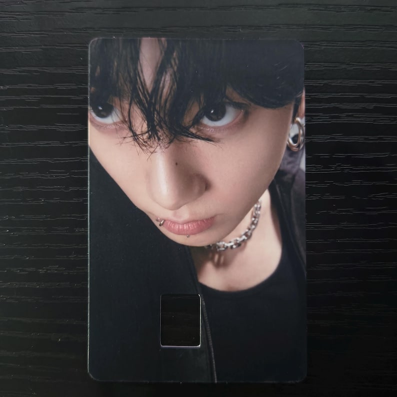 May include: A rectangular card featuring a close-up portrait of a person with dark hair and a piercing. The person is wearing a black top and a silver chain necklace. The card has a small rectangular cutout near the bottom.