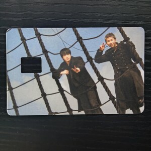 May include: A rectangular card featuring two people posing on a ship's rigging against a blue sky backdrop. The card has a black rectangular cutout. The people are wearing black outfits. The rigging is made of thick, dark ropes.