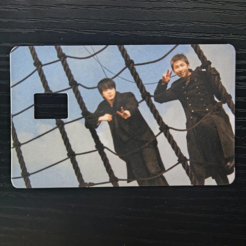 May include: A rectangular card featuring two people posing on a ship's rigging against a blue sky backdrop. The card has a black rectangular cutout. The people are wearing black outfits. The rigging is made of thick, dark ropes.