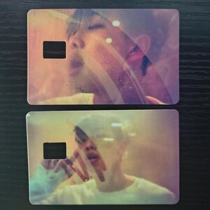 May include: Two rectangular cards with a blurred image of a person's face. The cards have a cutout square on the left side. The image has a soft, pastel color palette with pink, purple, and yellow hues. The background is a dark wood grain.