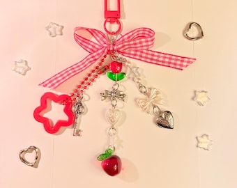 May include: A pink keychain with a red gingham ribbon, a red flower-shaped charm, and various dangling charms. Charms include a key, a cross, a bow, a heart-shaped locket, and a red apple. The keychain has a red clasp.
