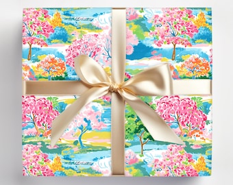 May include: A gift box wrapped in colorful floral patterned paper with a gold satin ribbon and bow. The paper features a landscape design with pink, orange, and green trees, blue water, and a yellow sky.
