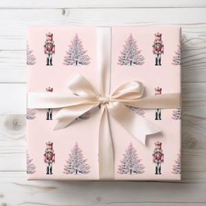 Puede incluir: Un regalo envuelto con un fondo rosa claro que presenta un patrón repetido de árboles de Navidad y cascanueces. El regalo está atado con una cinta y un lazo de satén color crema. El diseño del papel de regalo tiene un tema festivo.