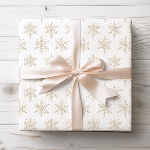 May include: A wrapped gift with white paper featuring a gold snowflake pattern. A light peach satin ribbon is tied in a bow around the package. The gift is presented on a white wooden surface.