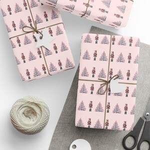 May include: Wrapped gifts with pink paper patterned with nutcrackers and Christmas trees. The gifts are tied with twine and have white tags. A ball of twine and scissors are also visible, suggesting a festive theme.