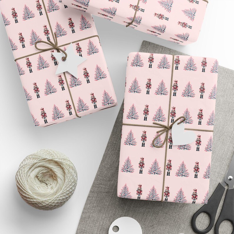 May include: Wrapped gifts with pink paper patterned with nutcrackers and Christmas trees. The gifts are tied with twine and have white tags. A ball of twine and scissors are also visible, suggesting a festive theme.