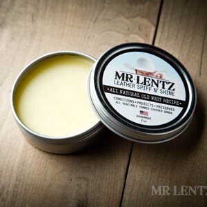 May include: A silver tin with a yellow label that reads "MR. LENTZ Leather Spiff N' Shine - All Natural Old West Recipe - Conditions, Protects, Preserves - All Vegetable Tanned Leather Goods - Handmade - 2 oz". The tin is open and a small amount of yellow balm is visible inside.