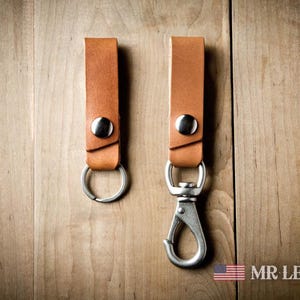 Pode incluir: Duas tiras de couro castanho-claro com ferragens de metal prateado. Uma tira possui um anel circular, enquanto a outra inclui um mosquet&atilde;o girat&oacute;rio. Ambas s&atilde;o fixadas com um bot&atilde;o de press&atilde;o prateado. O texto diz MR LENTZ.com.