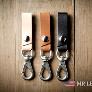 Pode incluir: Tr&ecirc;s tiras de couro para chaves nas cores castanho claro, castanho m&eacute;dio e preto. Cada tira possui um bot&atilde;o de press&atilde;o de metal prateado e um gancho mosquet&atilde;o girat&oacute;rio. O texto MR LENTZ.com e uma pequena bandeira americana s&atilde;o vis&iacute;veis.