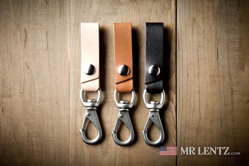 Pode incluir: Tr&ecirc;s tiras de couro para chaves nas cores castanho claro, castanho m&eacute;dio e preto. Cada tira possui um bot&atilde;o de press&atilde;o de metal prateado e um gancho mosquet&atilde;o girat&oacute;rio. O texto MR LENTZ.com e uma pequena bandeira americana s&atilde;o vis&iacute;veis.