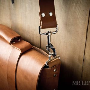 Pode incluir: Uma bolsa de mensageiro de couro marrom com um fecho de prata e uma al&ccedil;a. A bolsa &eacute; feita de couro de alta qualidade e tem um design cl&aacute;ssico.