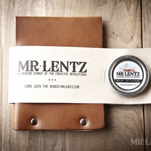 May include: A brown leather notebook with a small tin of leather polish. The tin has a label that says "MR LENTZ Leather Spoff N Shine" and "Natural and Handmade". The notebook has a label that says "MR LENTZ" and "A modern cowboy of the creative revolution".