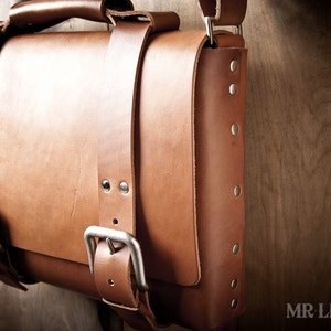 Pode incluir: Uma bolsa de mensageiro de couro marrom com fecho de aba e v&aacute;rias fivelas. A bolsa tem uma al&ccedil;a de ombro e &eacute; decorada com rebites prateados.