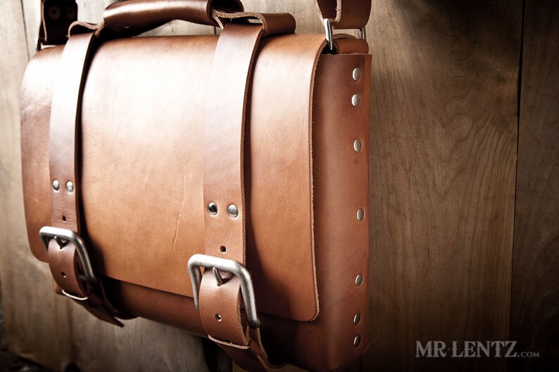 Pode incluir: Uma bolsa de mensageiro de couro marrom com fecho de aba e v&aacute;rias fivelas. A bolsa tem uma al&ccedil;a de ombro e &eacute; decorada com rebites prateados.