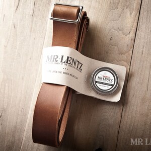 Pode incluir: Uma tira de couro marrom com uma fivela de prata e uma pequena lata de condicionador de couro. A lata tem o texto "MR. LENTZ" e "Leather Balm & Soap". A tira &eacute; embalada com um r&oacute;tulo branco que diz "MR. LENTZ" e "The Creative Rebellion".