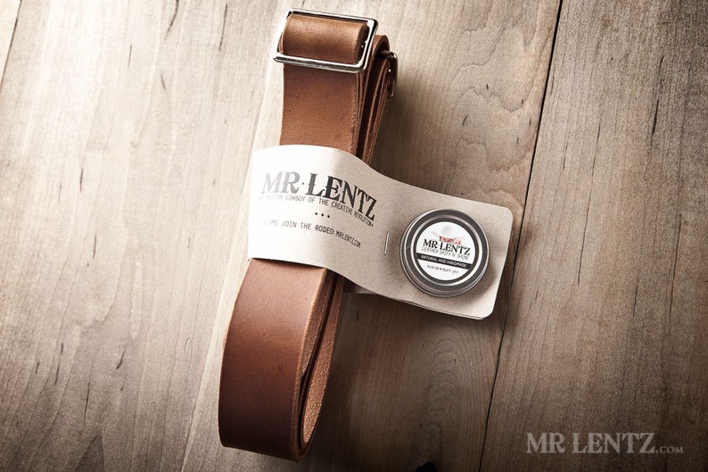 Pode incluir: Uma tira de couro marrom com uma fivela de prata e uma pequena lata de condicionador de couro. A lata tem o texto "MR. LENTZ" e "Leather Balm & Soap". A tira &eacute; embalada com um r&oacute;tulo branco que diz "MR. LENTZ" e "The Creative Rebellion".