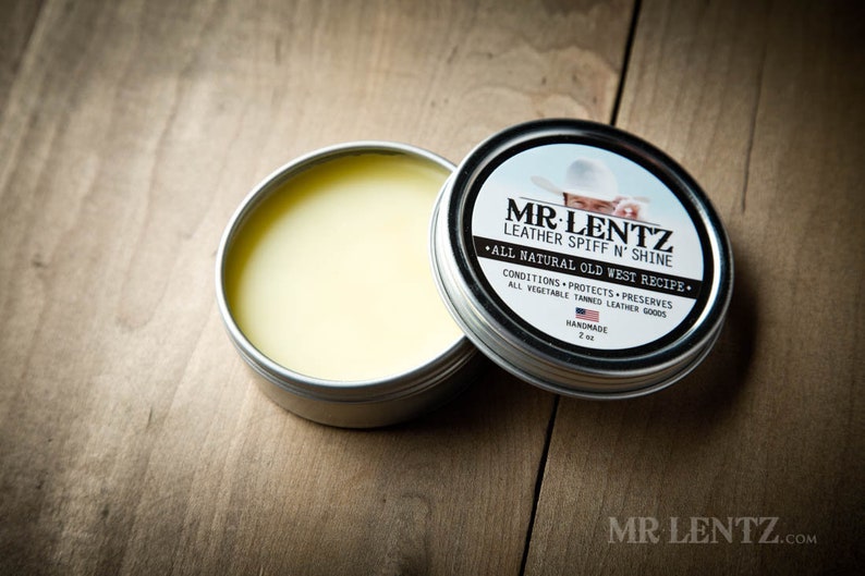 May include: A silver tin with a yellow label that reads "MR. LENTZ Leather Spiff N' Shine - All Natural Old West Recipe - Conditions, Protects, Preserves - All Vegetable Tanned Leather Goods - Handmade - 2 oz". The tin is open and a small amount of yellow balm is visible inside.