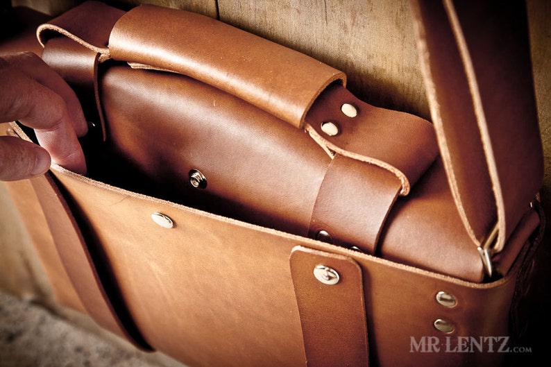 Pode incluir: Uma bolsa de couro marrom com fecho de aba e uma al&ccedil;a longa. A bolsa tem v&aacute;rios rebites de metal prateado e est&aacute; aberta para revelar o interior.