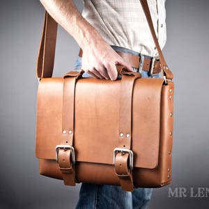 Pode incluir: Uma bolsa de mensageiro de couro marrom com duas fivelas e uma al&ccedil;a longa. A bolsa tem um fecho de aba e &eacute; feita de couro de alta qualidade.