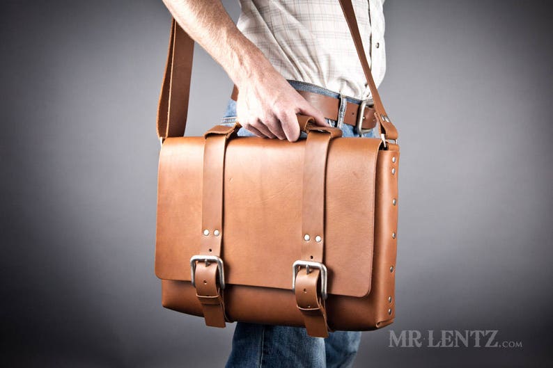 Pode incluir: Uma bolsa de mensageiro de couro marrom com duas fivelas e uma al&ccedil;a longa. A bolsa tem um fecho de aba e &eacute; feita de couro de alta qualidade.