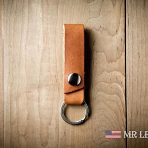 Pode incluir: Um porta-chaves de couro castanho-claro com uma tira retangular dobrada sobre um bot&atilde;o de press&atilde;o de metal prateado e um anel circular. O texto MR LENTZ.com est&aacute; impresso no canto inferior direito junto a um &iacute;cone da bandeira americana.