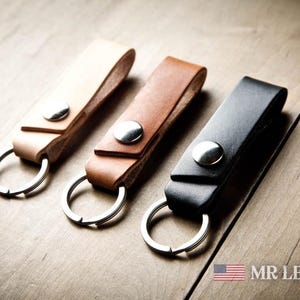 Pode incluir: Tr&ecirc;s porta-chaves de couro em bege claro, castanho m&eacute;dio e preto, cada um com um bot&atilde;o de press&atilde;o de metal prateado e uma argola circular. O texto MR LENTZ.com com um pequeno &iacute;cone da bandeira americana &eacute; vis&iacute;vel no canto inferior.