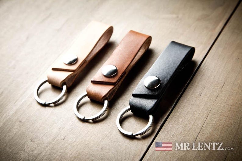 Pode incluir: Tr&ecirc;s porta-chaves de couro em bege claro, castanho m&eacute;dio e preto, cada um com um bot&atilde;o de press&atilde;o de metal prateado e uma argola circular. O texto MR LENTZ.com com um pequeno &iacute;cone da bandeira americana &eacute; vis&iacute;vel no canto inferior.