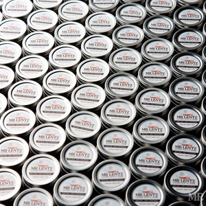 May include: A close-up view of a large quantity of small, silver, round tins with white labels that read "MR. LENTZ" in black lettering. The tins are arranged in a grid pattern.
