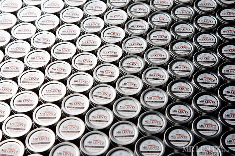 May include: A close-up view of a large quantity of small, silver, round tins with white labels that read "MR. LENTZ" in black lettering. The tins are arranged in a grid pattern.